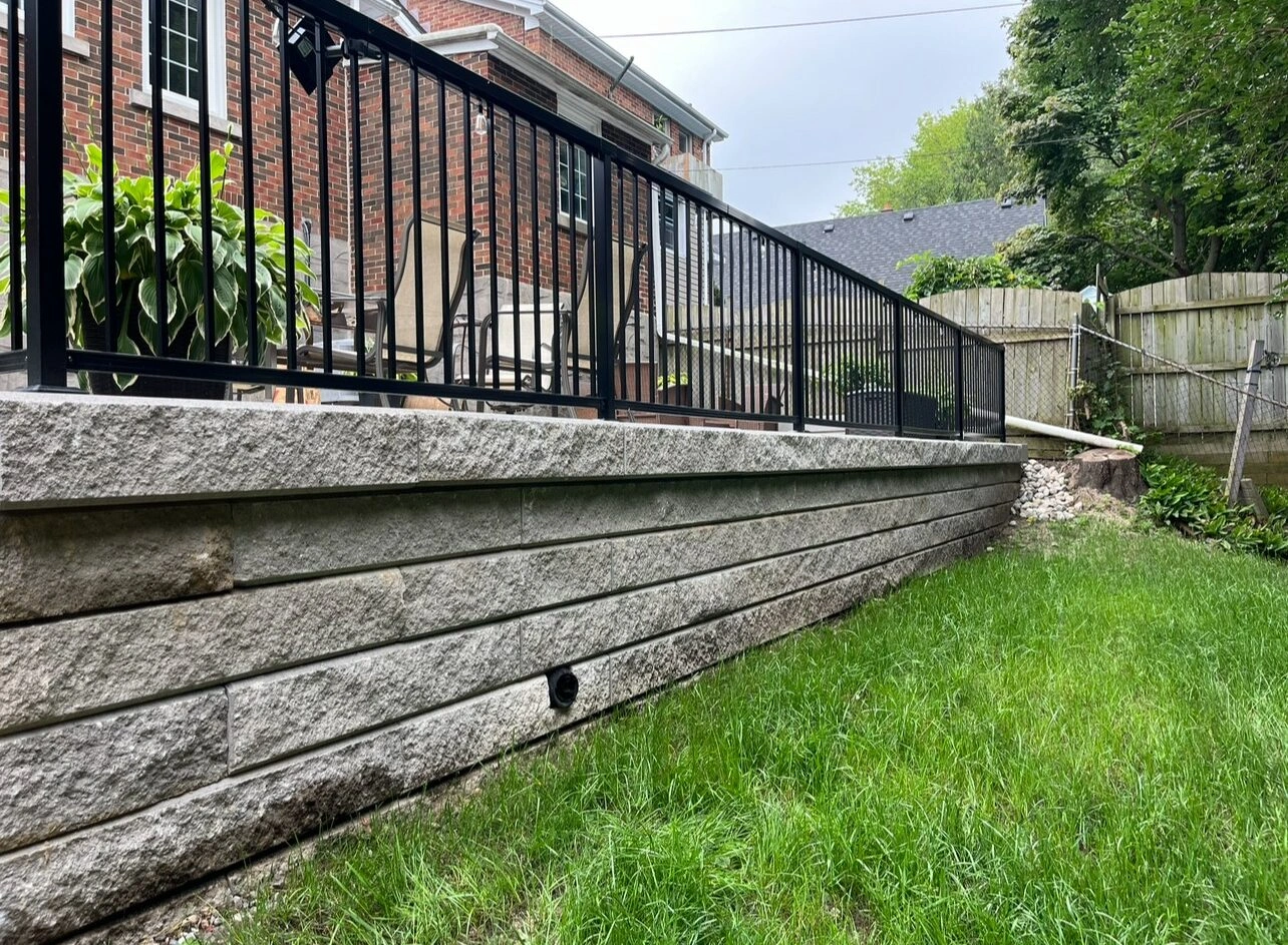 Brick house with elevated stone patio, black metal railing, surrounded by lush greenery and fenced backyard. Outdoor seating area visible in background.