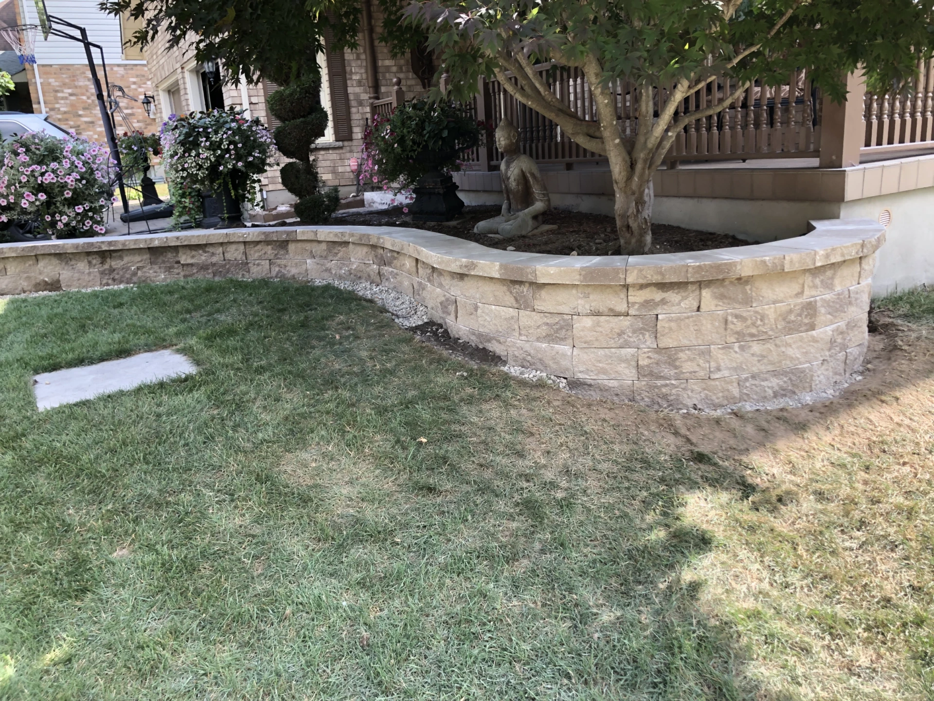 A garden features a curved stone retaining wall, vibrant flowers, a seated sculpture beneath a tree, and a nearby porch.