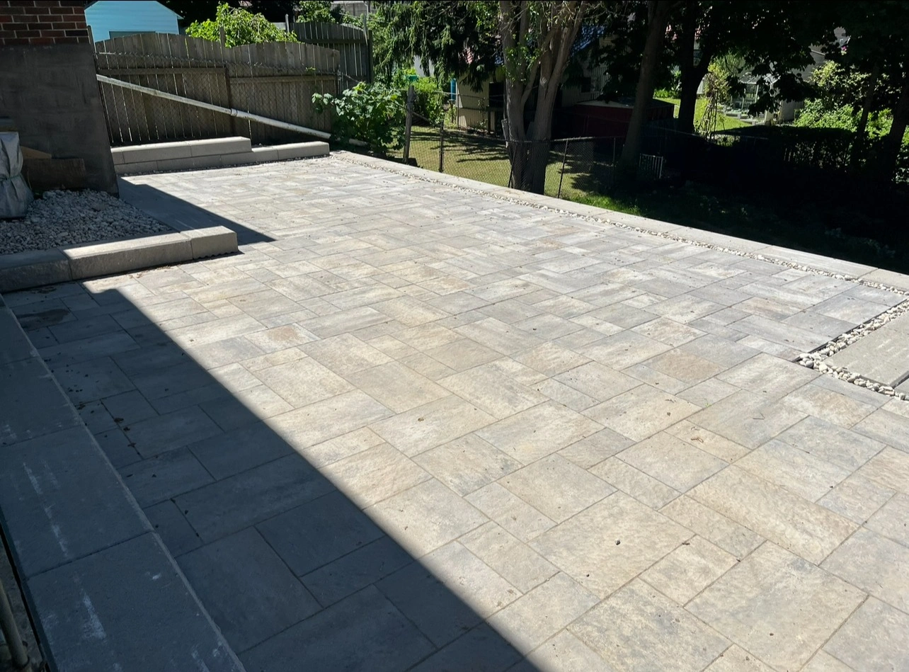 A newly paved patio with light grey tiles, bordered by a fence and trees, under bright sunlight and partial shadow.