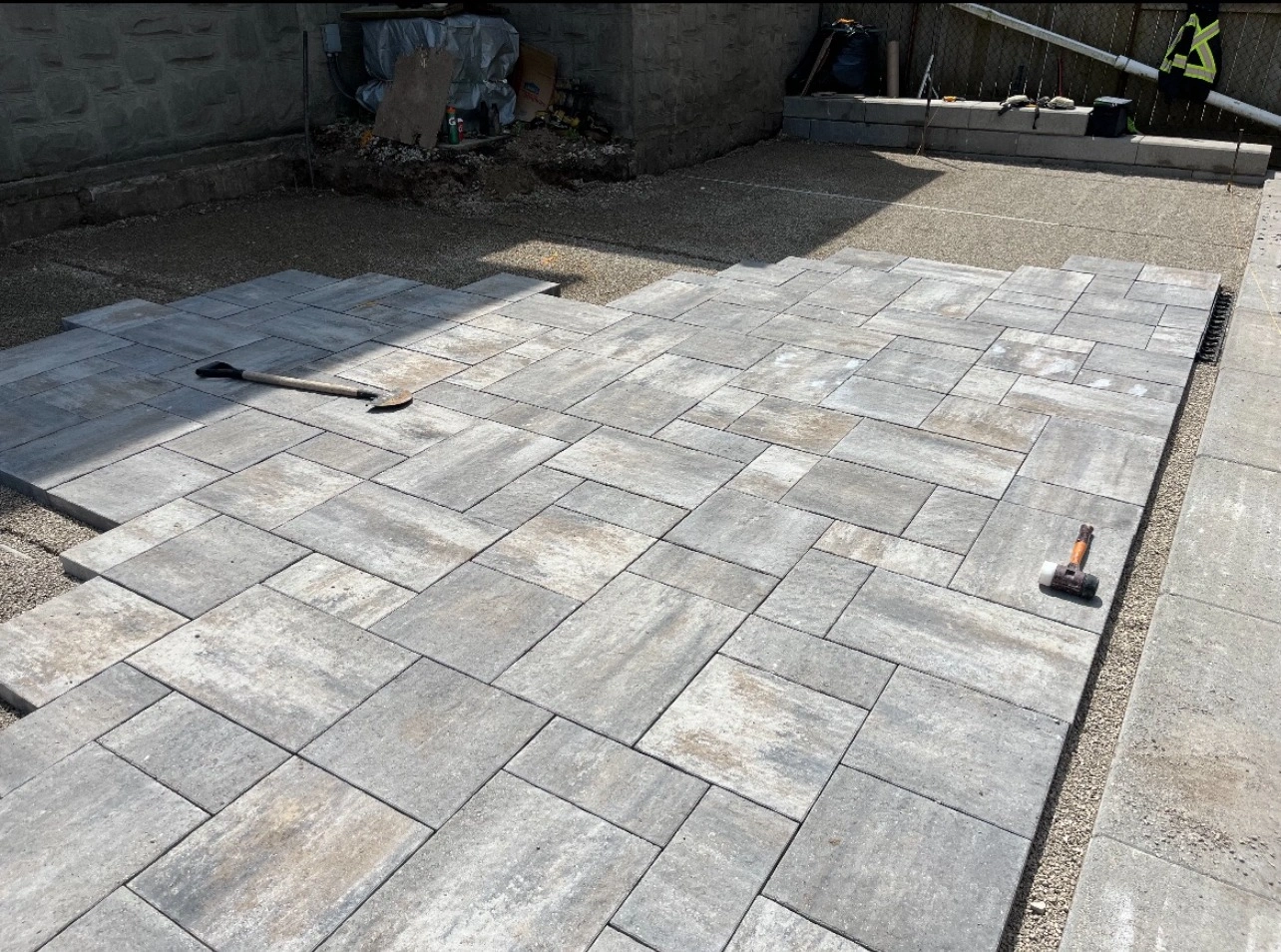 A paved area under construction with interlocking stones; a hammer and tools are visible. A person wearing a construction vest is nearby.