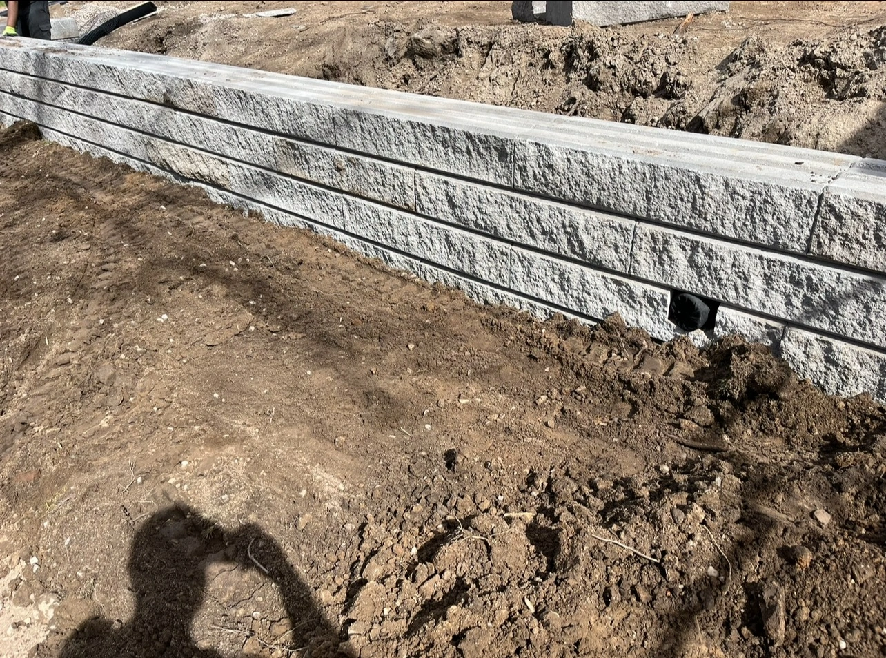 Stone retaining wall under construction on a dirt site, with a person’s shadow visible in the foreground.