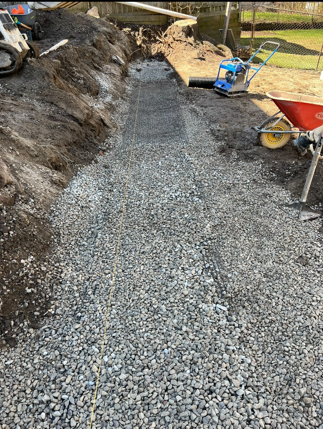 A gravel path under construction with tools and equipment scattered around, including a wheelbarrow, compactor, and string line.