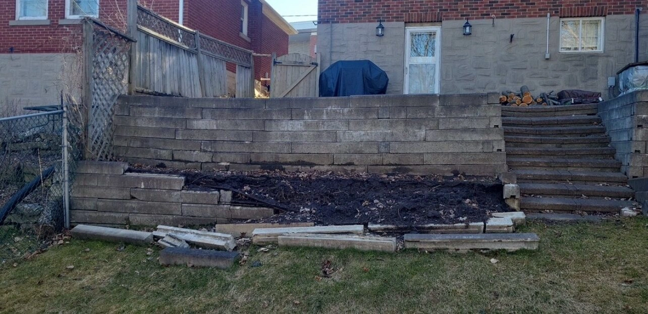A brick house with a damaged retaining wall, wooden steps, and yard debris, seen in a residential neighborhood setting.