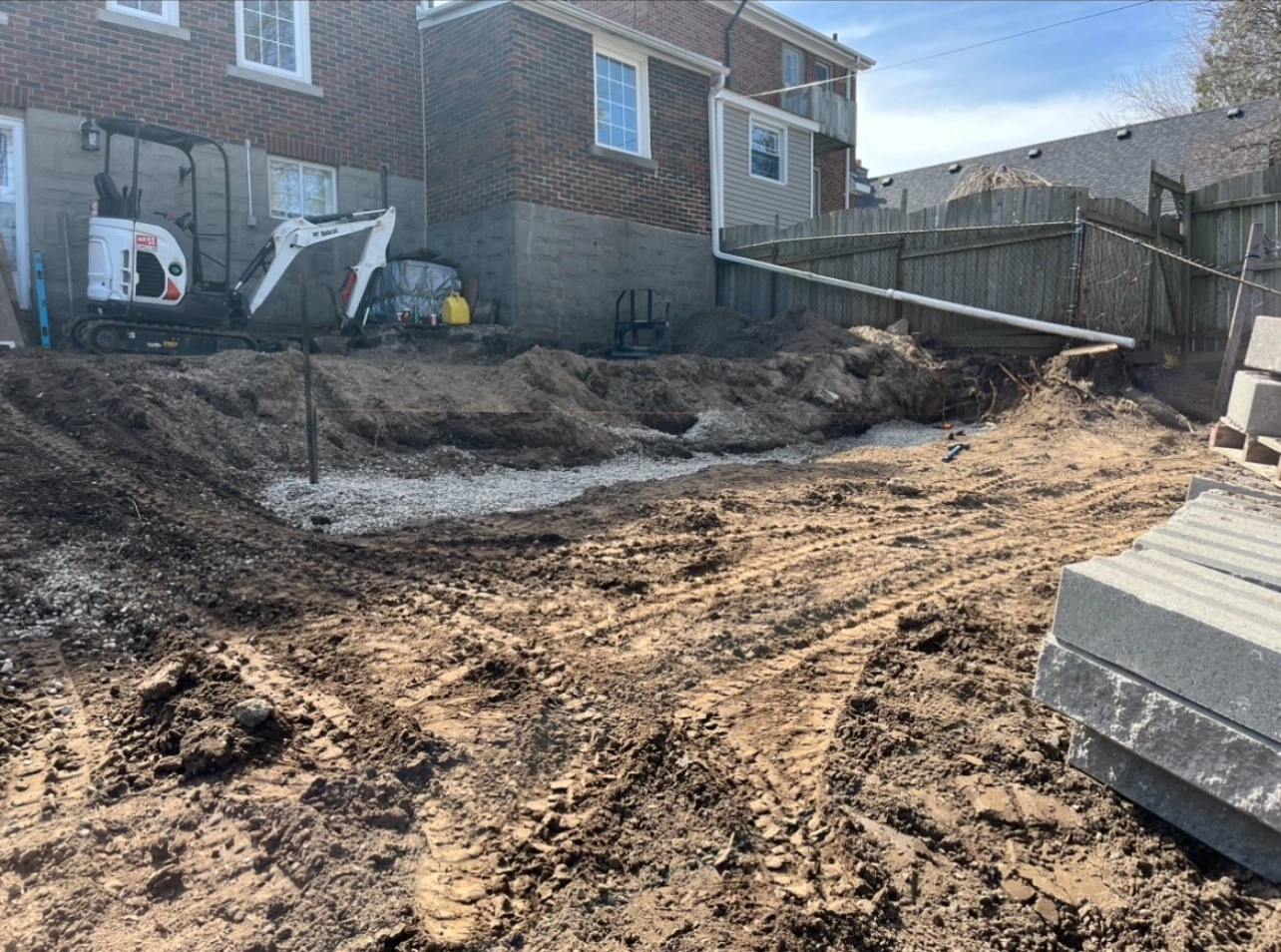 A construction site with excavated dirt, a small excavator, and a brick house in the background. Pallets of materials are visible.