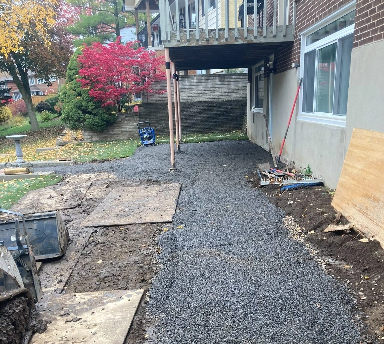 A backyard construction scene features gravel path under a raised balcony, surrounded by colorful autumn trees. No landmarks or historical buildings visible.