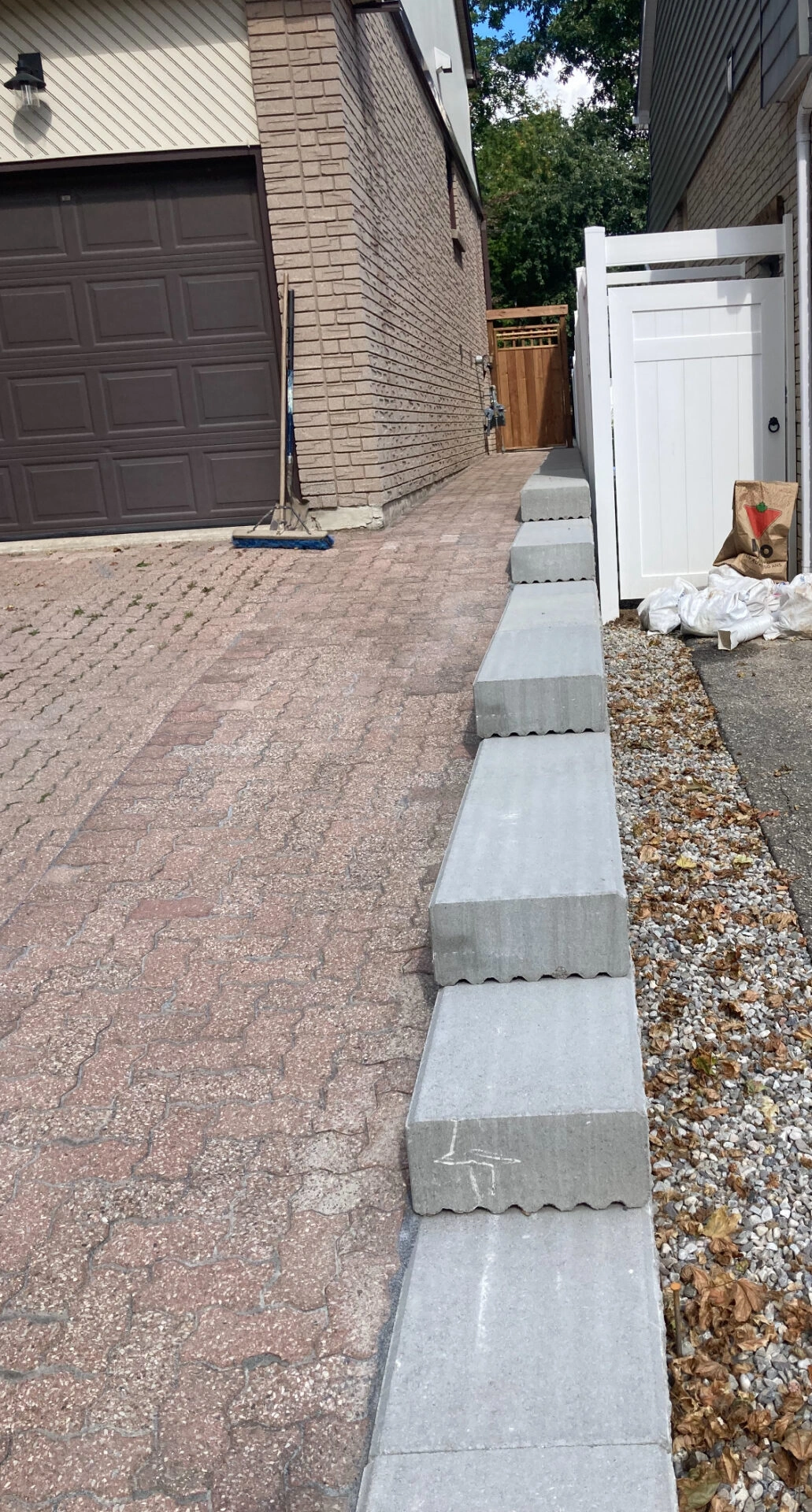 Brick driveway next to a house with a garage, bordered by stacked concrete blocks and a white fence, scattered leaves nearby.