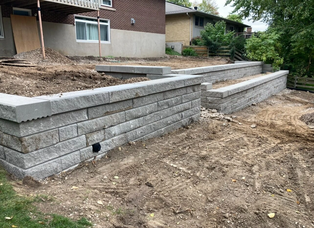 The image shows a partially constructed retaining wall in a residential backyard, featuring stacked stone blocks and surrounding soil, with trees nearby.