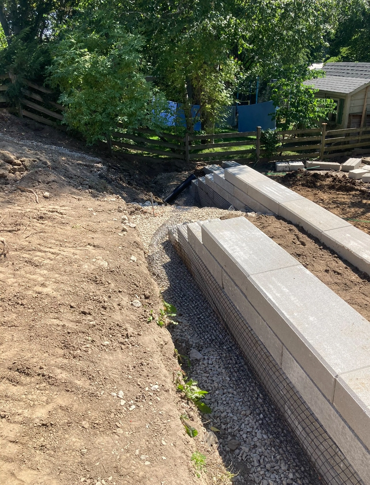 A newly constructed stone retaining wall with gravel foundation, surrounded by dirt, trees, and a wooden fence in a backyard setting.