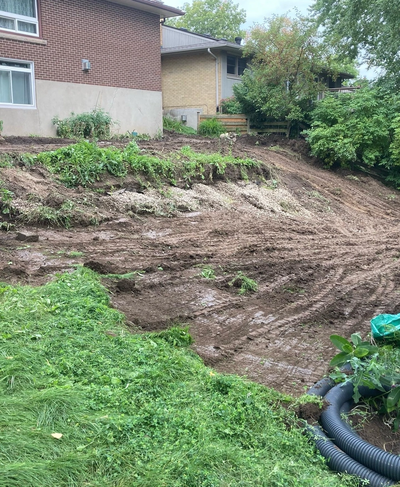 A sloped backyard with fresh dirt, tire tracks, and scattered greenery. A brick house is visible. No people or landmarks are present.