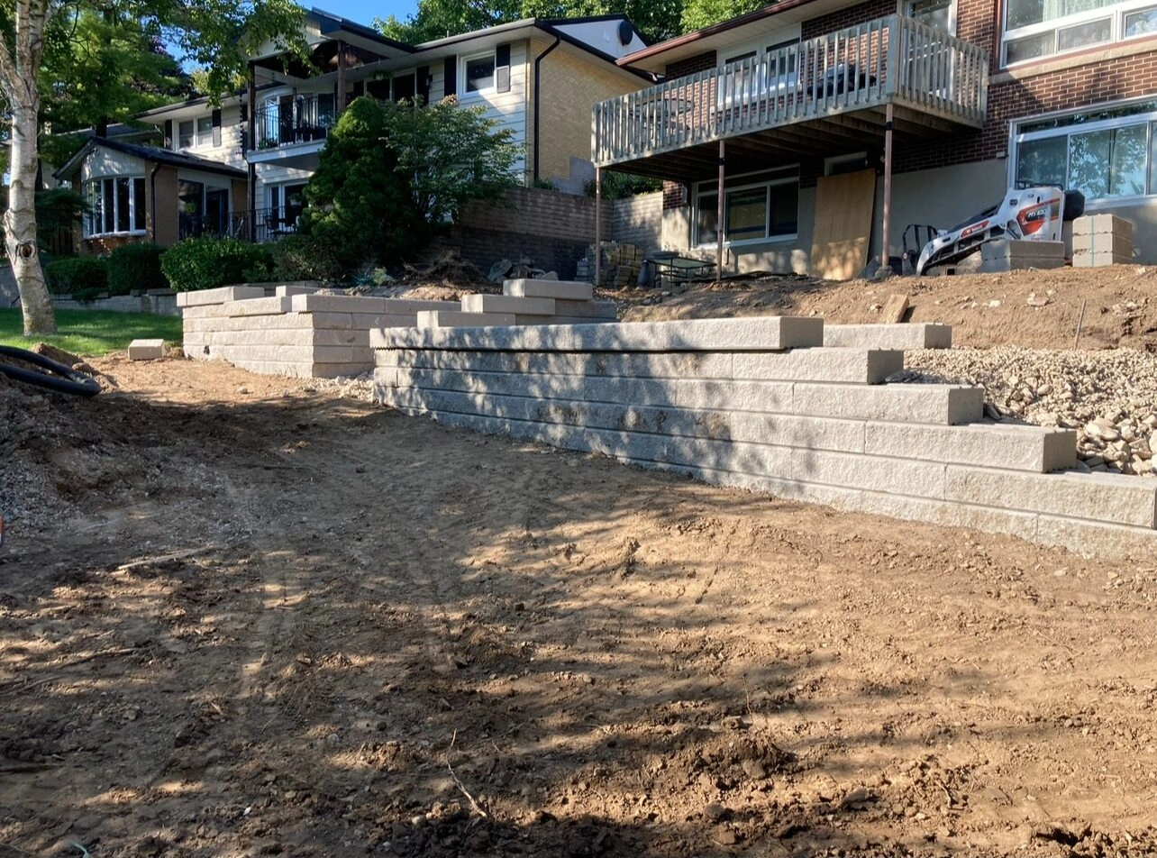 Newly constructed stone retaining wall in a residential backyard, with modern houses in the background and surrounding greenery. No landmarks visible.