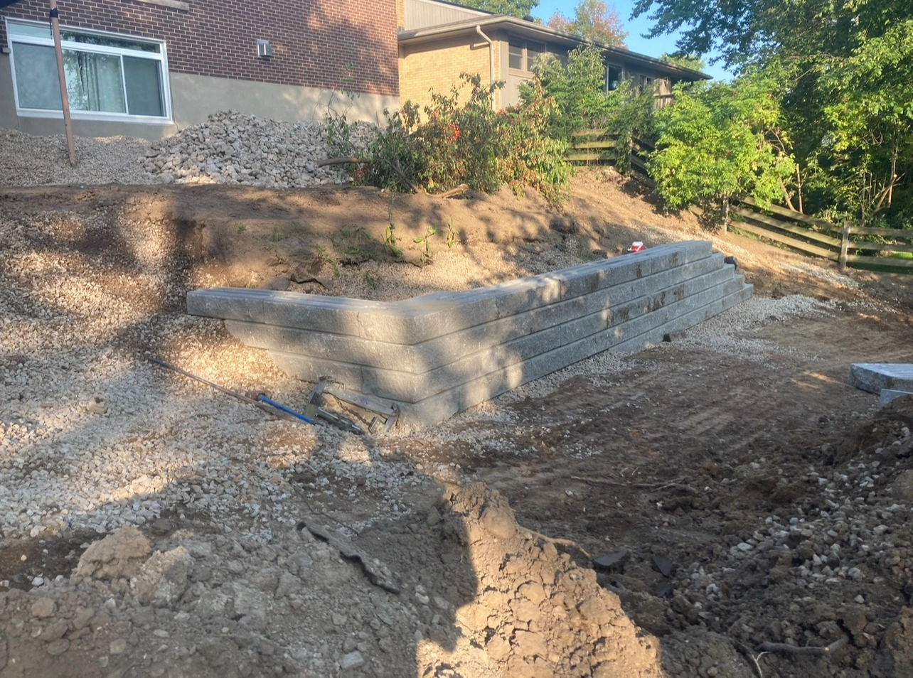A partially constructed retaining wall made of concrete blocks sits beside a residential brick house with tools and gravel around.