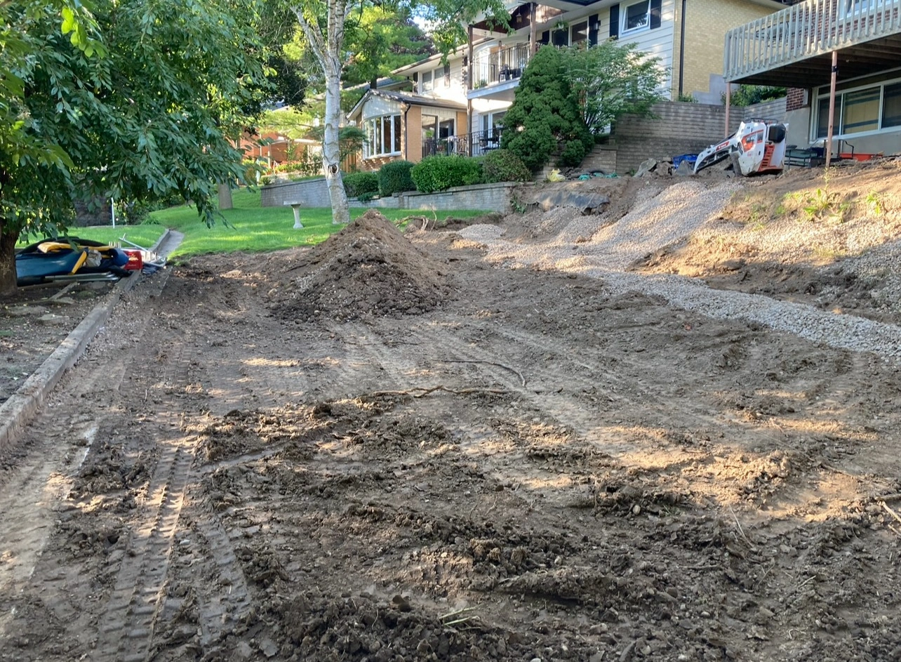 A residential backyard undergoing landscaping with piles of dirt and gravel. A small construction vehicle is parked near a tree and houses.