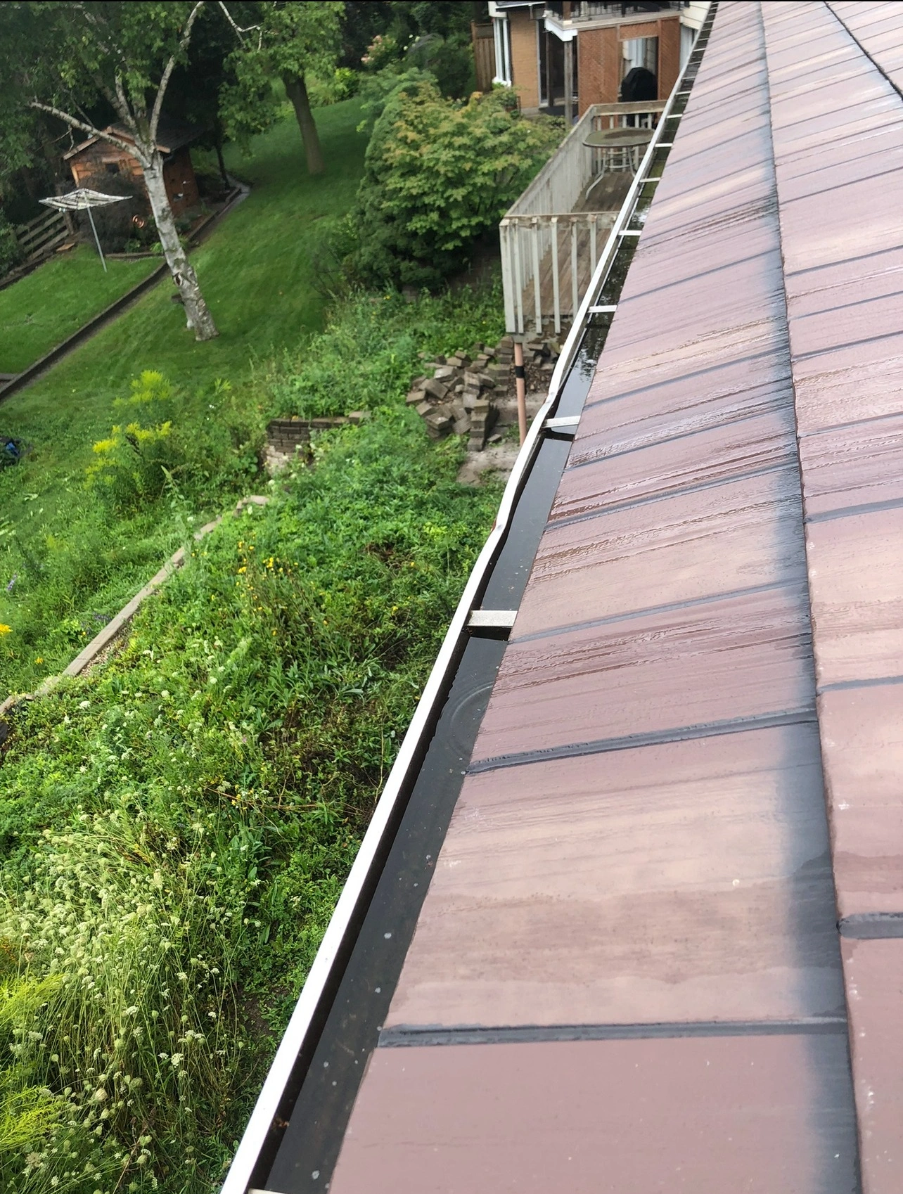 Rooftop view of a lush garden with trees and a small shed, alongside a house's deck and tiled roof.