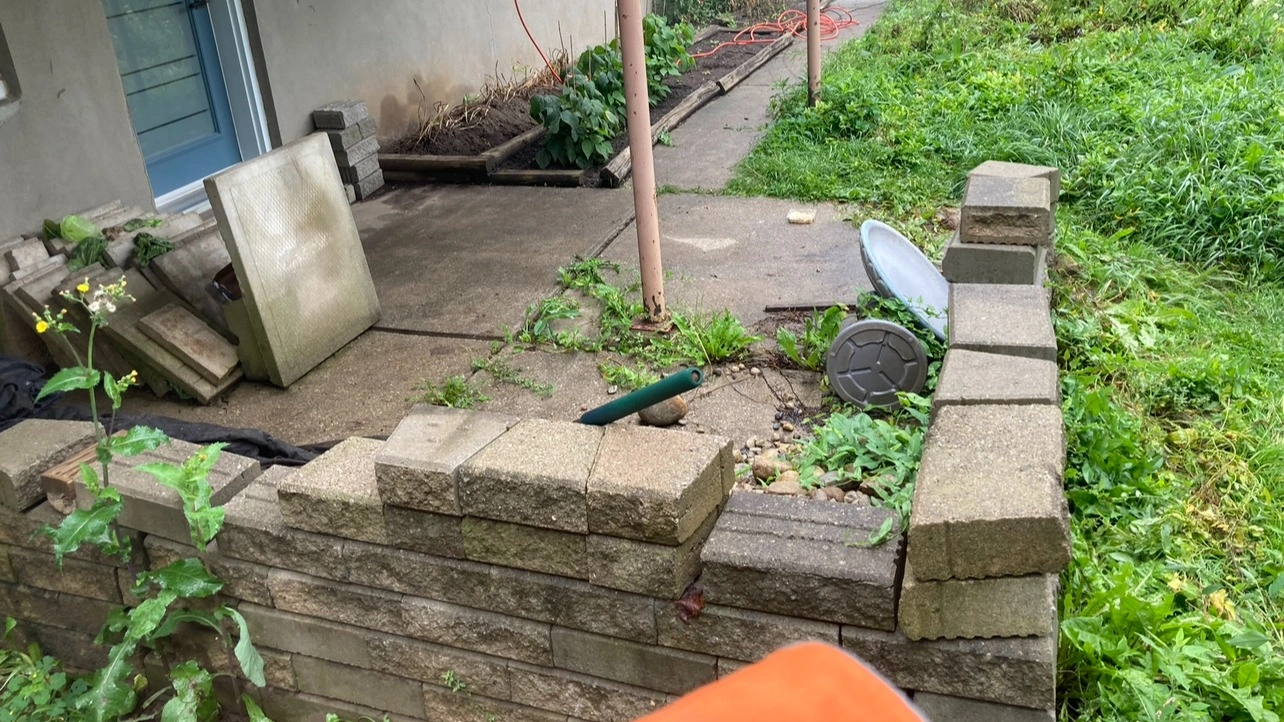 A small patio with bricks and tools is surrounded by greenery. A pathway leads to a garden area with scattered equipment.