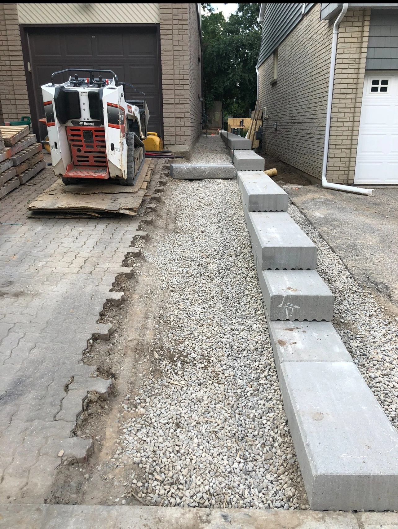 A construction site between two buildings shows a Bobcat loader, paving stones, gravel, and stacked concrete blocks.