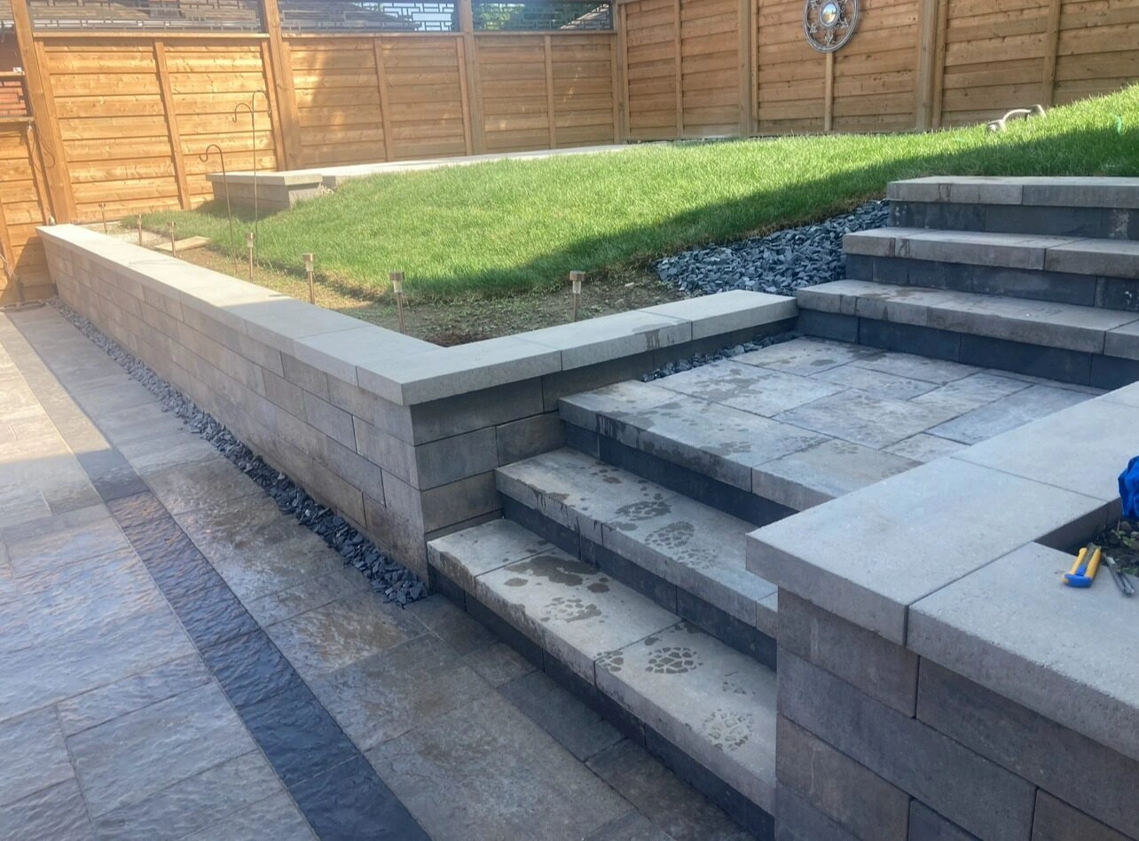 Stone steps lead to the grassy area in a fenced backyard. Tools are scattered on a concrete wall.