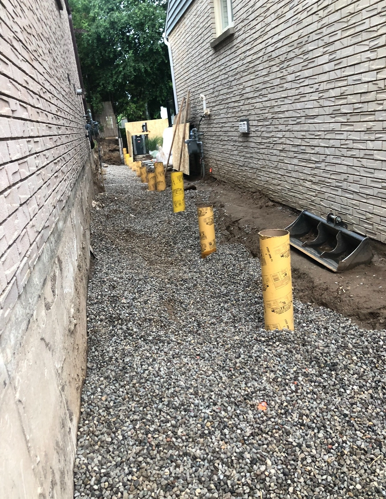 Narrow construction site between two brick buildings with gravel ground and yellow concrete form tubes. Equipment and materials are scattered around.