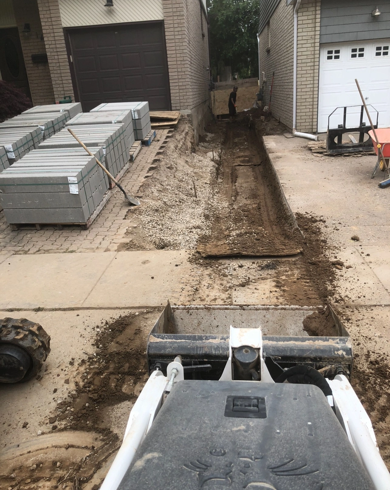 A construction vehicle digs a trench between two brick garages; a person stands at the end, surrounded by stacked materials and tools.