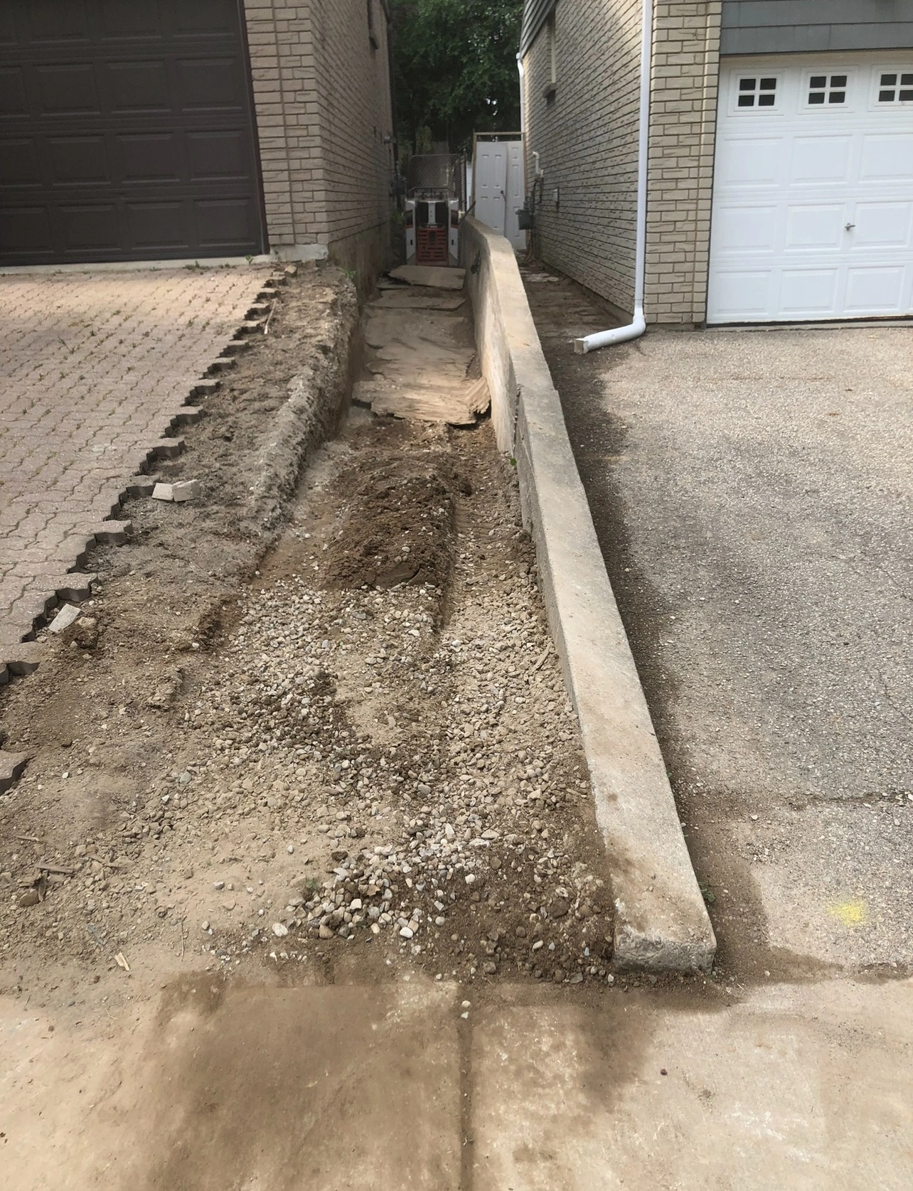 Narrow alleyway between two brick houses shows a partially excavated trench filled with dirt and gravel, leading to a small gate in the background.