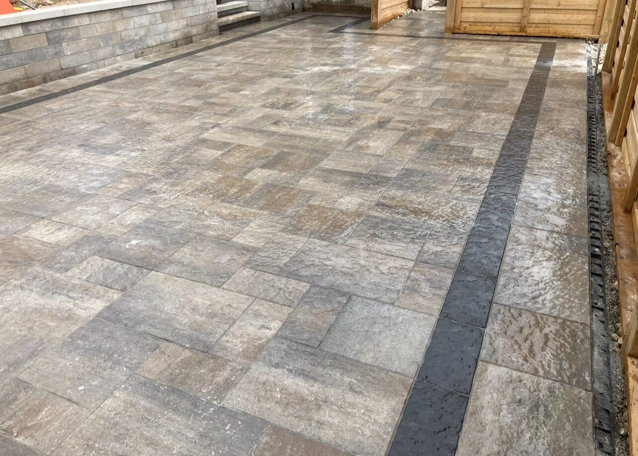 Wet, patterned stone patio with dark borders, enclosed by wooden fencing and a stone retaining wall with small stairs leading up.