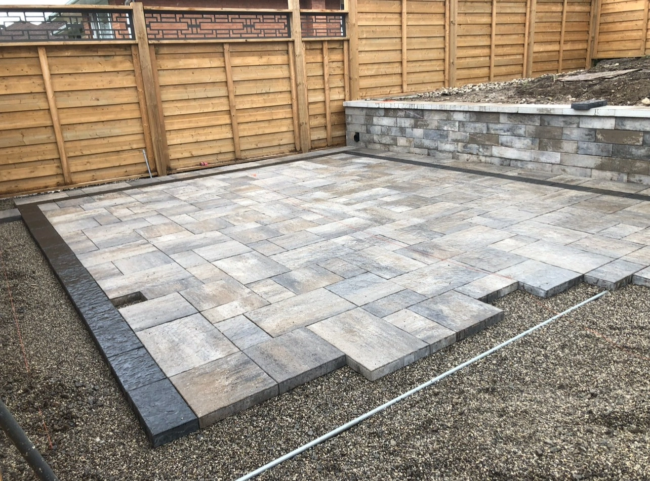 A neatly paved patio with interlocking stone design, enclosed by wooden fence and stone retaining wall, under construction.