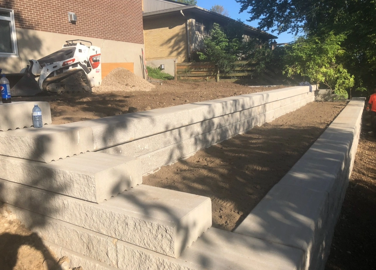 A construction site features a small excavator, retaining wall, and two water bottles on the ground, located between two brick buildings.