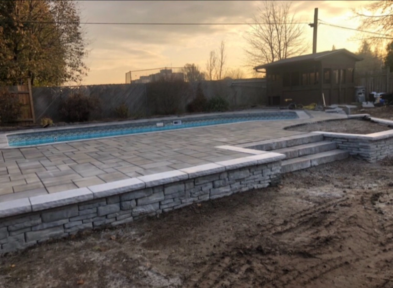 A stone patio with steps leads to a rectangular pool. Trees and a wooden structure surround the area against a sunset sky.