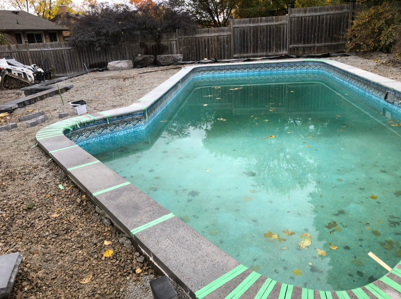 A partially constructed backyard pool with a gravel surround, some leaves in water, wooden fence, and construction equipment nearby.