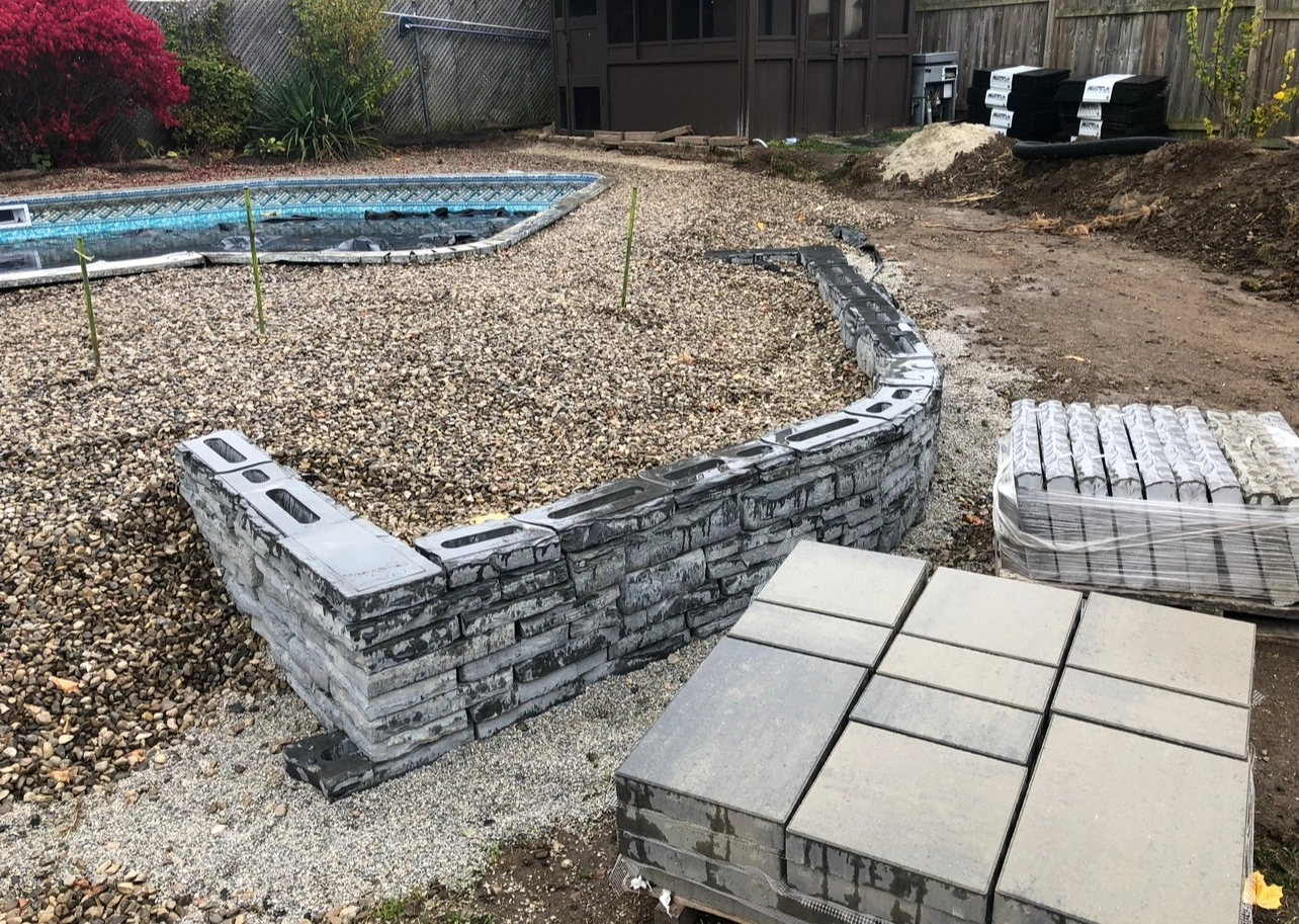 Backyard construction site with a partially built stone wall, gravel, paving stones, and an empty pool, surrounded by wooden fencing.