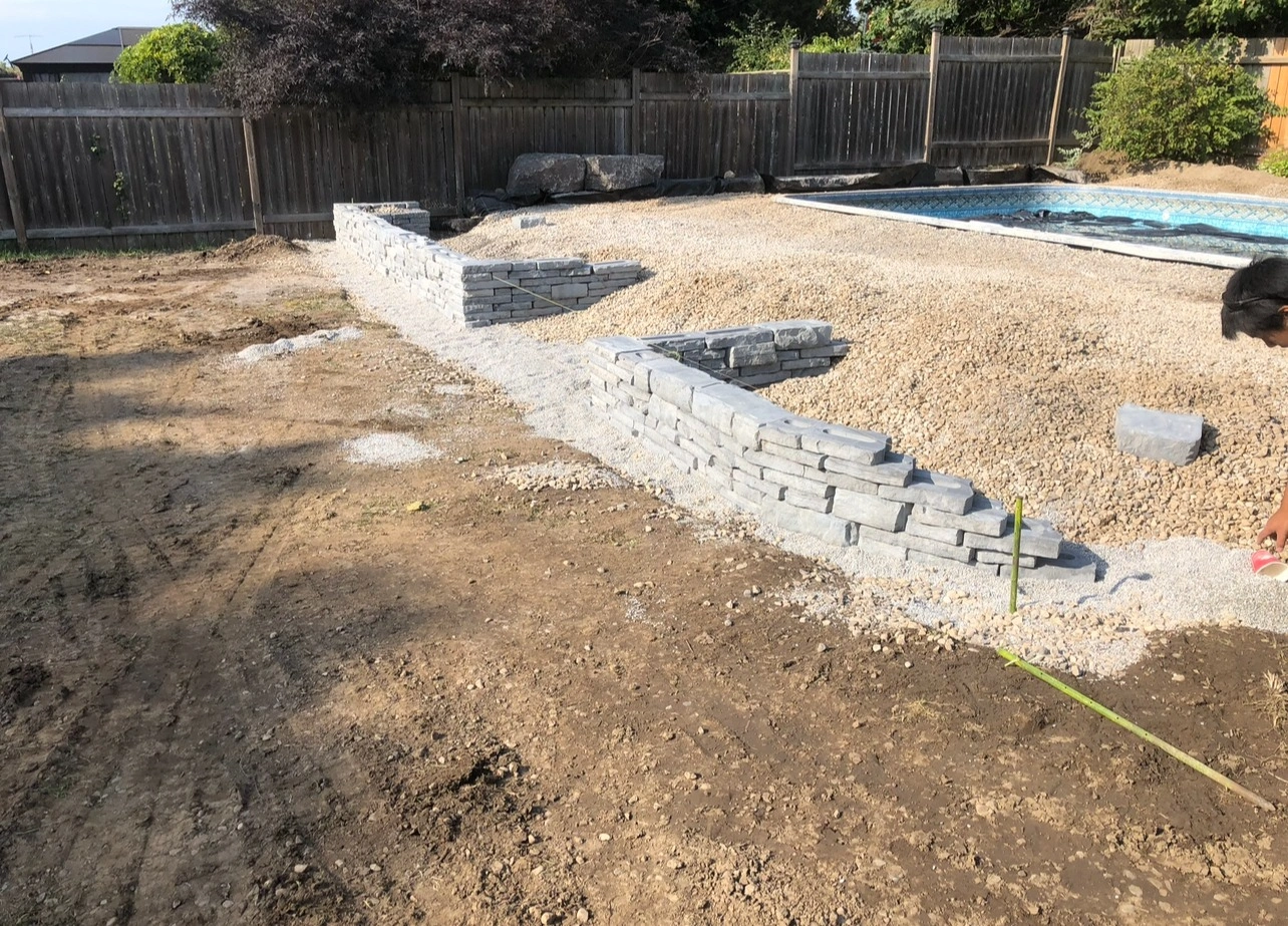 A person works on a rock wall near a fenced yard, with an adjacent gravel area and a covered pool.