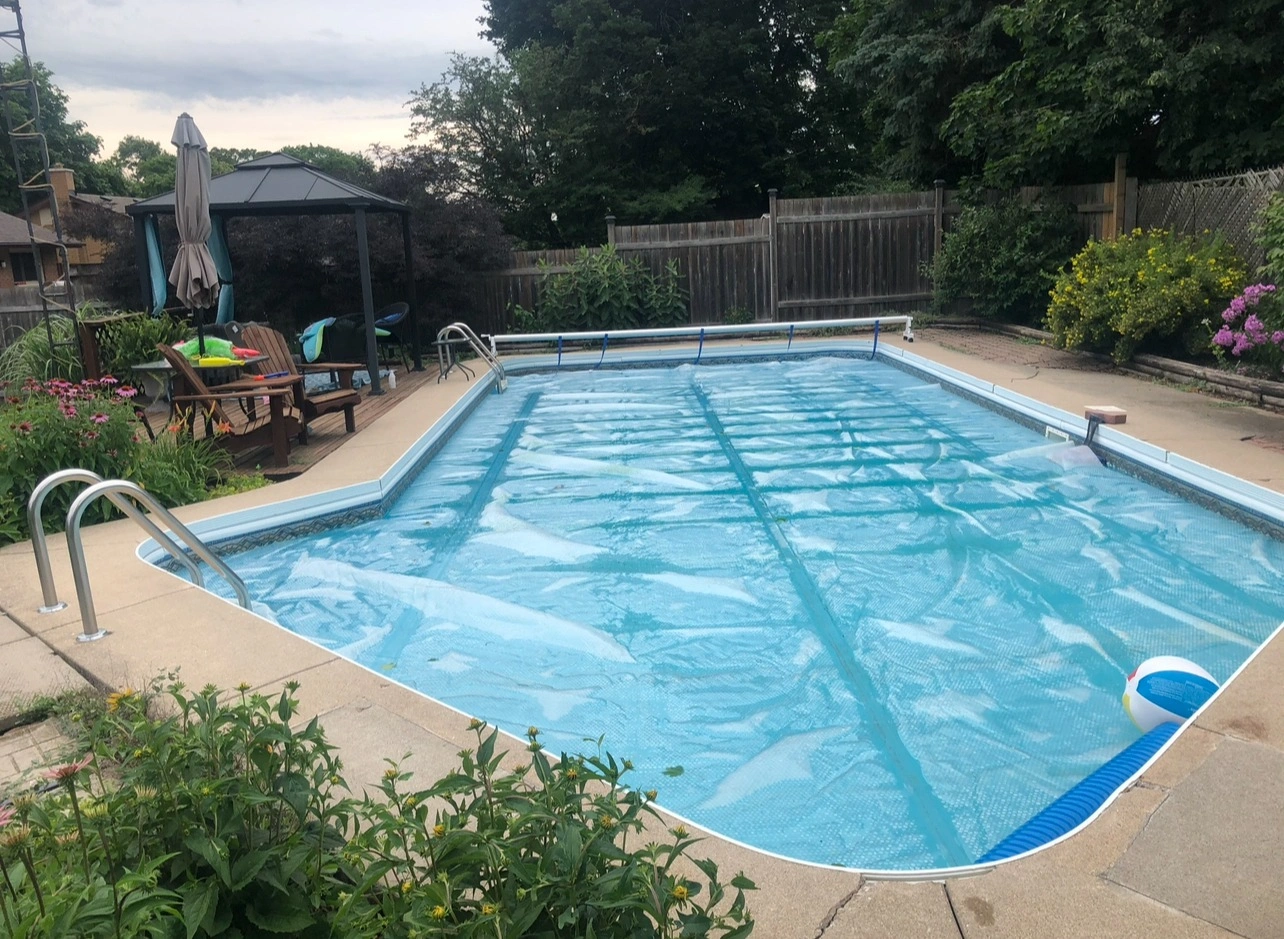 A backyard scene with a covered swimming pool, lounge chairs, umbrella, and garden. Fenced perimeter, lush greenery, and overcast skies.