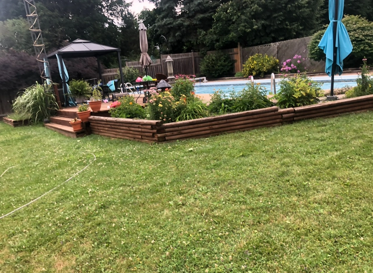 A backyard features a swimming pool, gazebo, umbrella, and lush greenery surrounded by wooden planters and a grassy lawn.