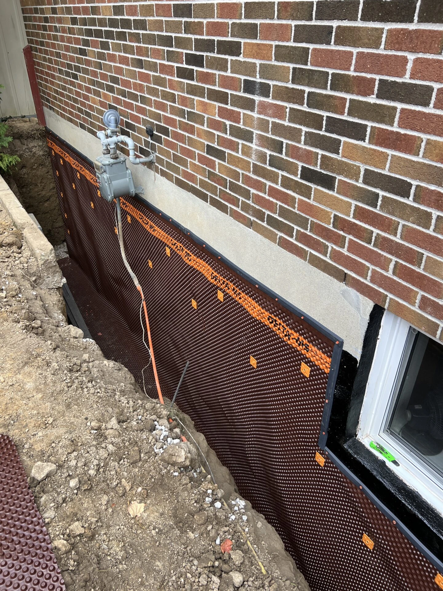 A brick building's foundation is being waterproofed with a drainage mat, visible gas meter, and window nearby, amid an excavation.