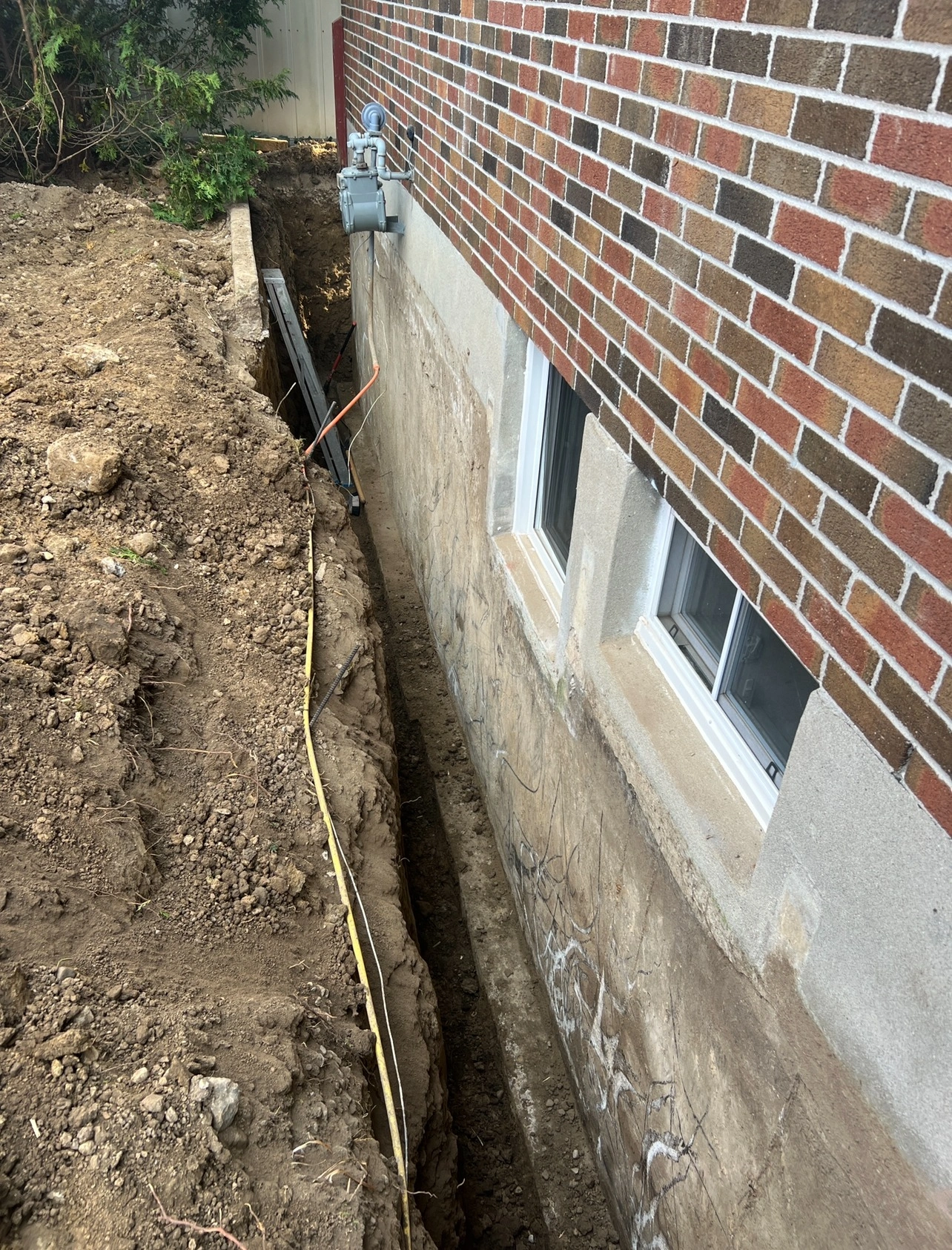 A trench is dug alongside a brick building foundation with exposed utility lines and unfinished excavation, indicating possible construction or repair work.