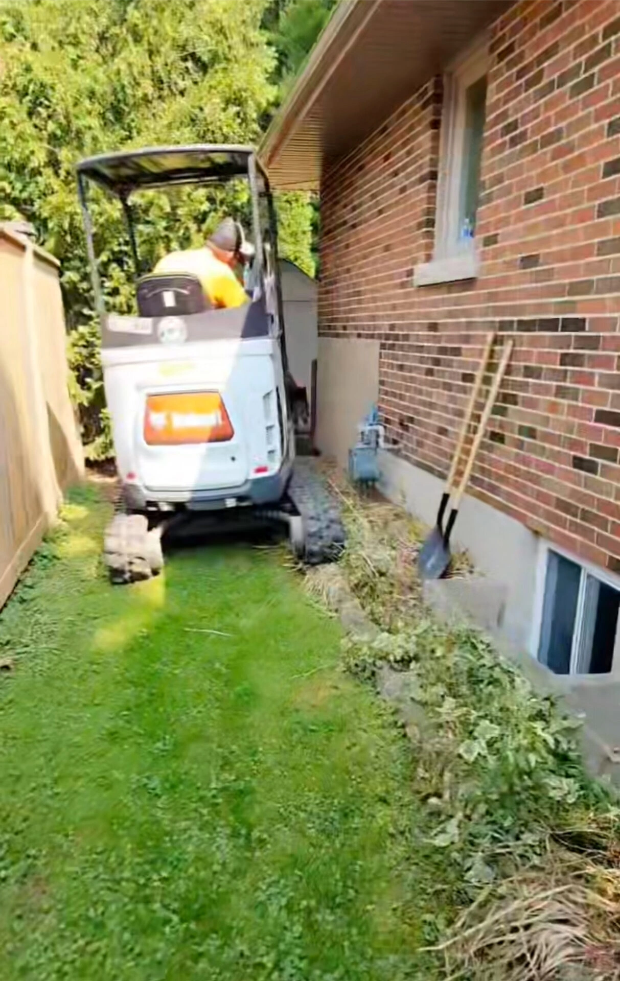 A person operates a small excavator beside a brick house with tools leaning against the wall, surrounded by grass and shrubs.