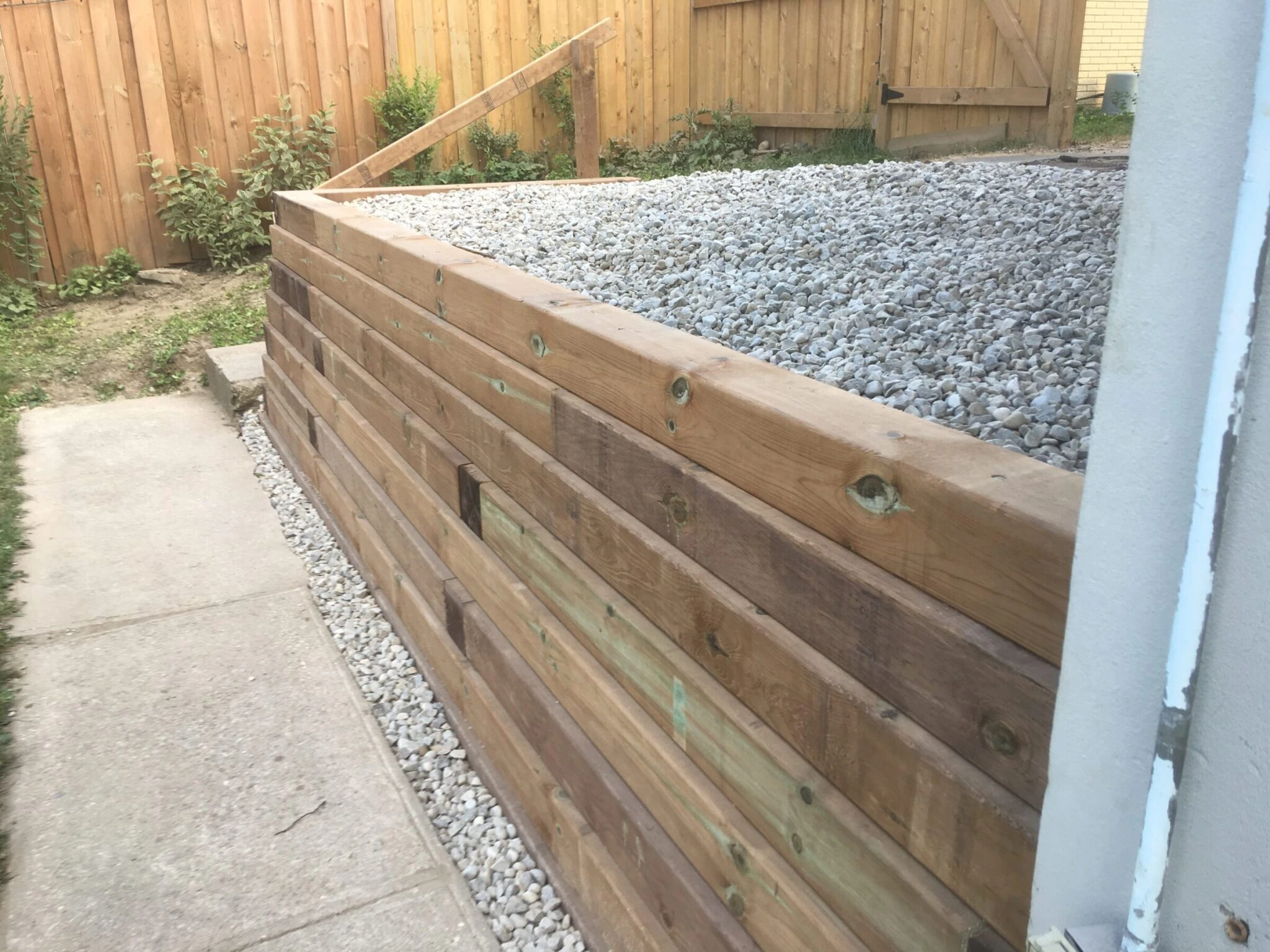A wooden retaining wall with gravel on top lines a narrow concrete pathway in a fenced backyard with sparse vegetation.