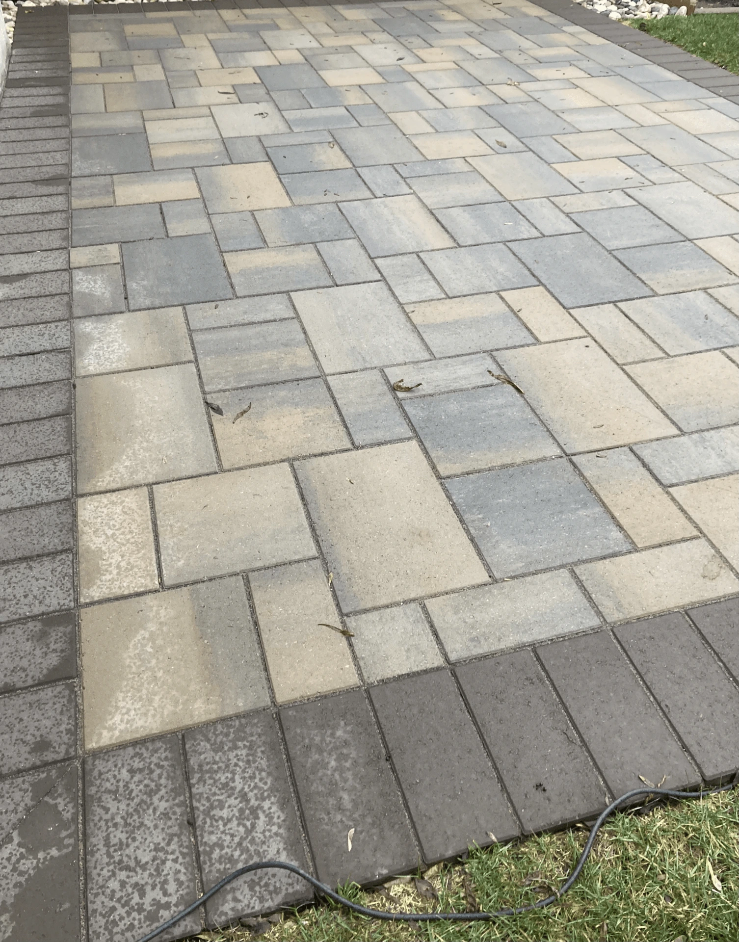 A neatly arranged patio features rectangular, gray and beige stone pavers with a darker border, surrounded by grass and scattered small leaves.