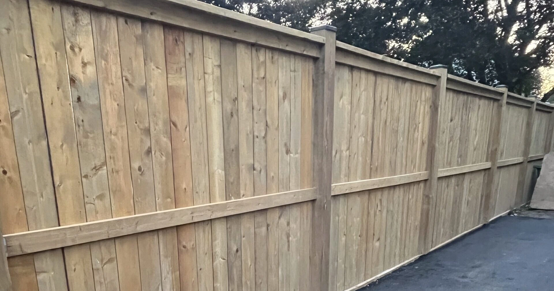 Wooden fence alongside an asphalt path, bordered by trees in a natural, outdoor setting. No people or landmarks are visible.