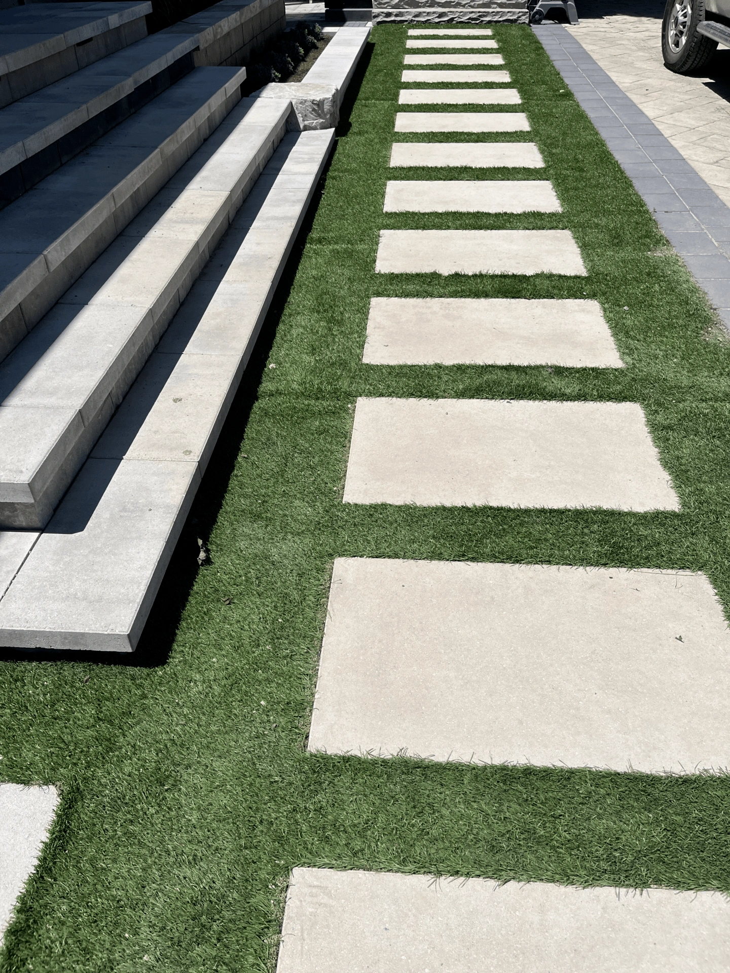 A pathway of rectangular stone slabs on green grass leads to concrete steps and a parked vehicle, under bright sunlight.