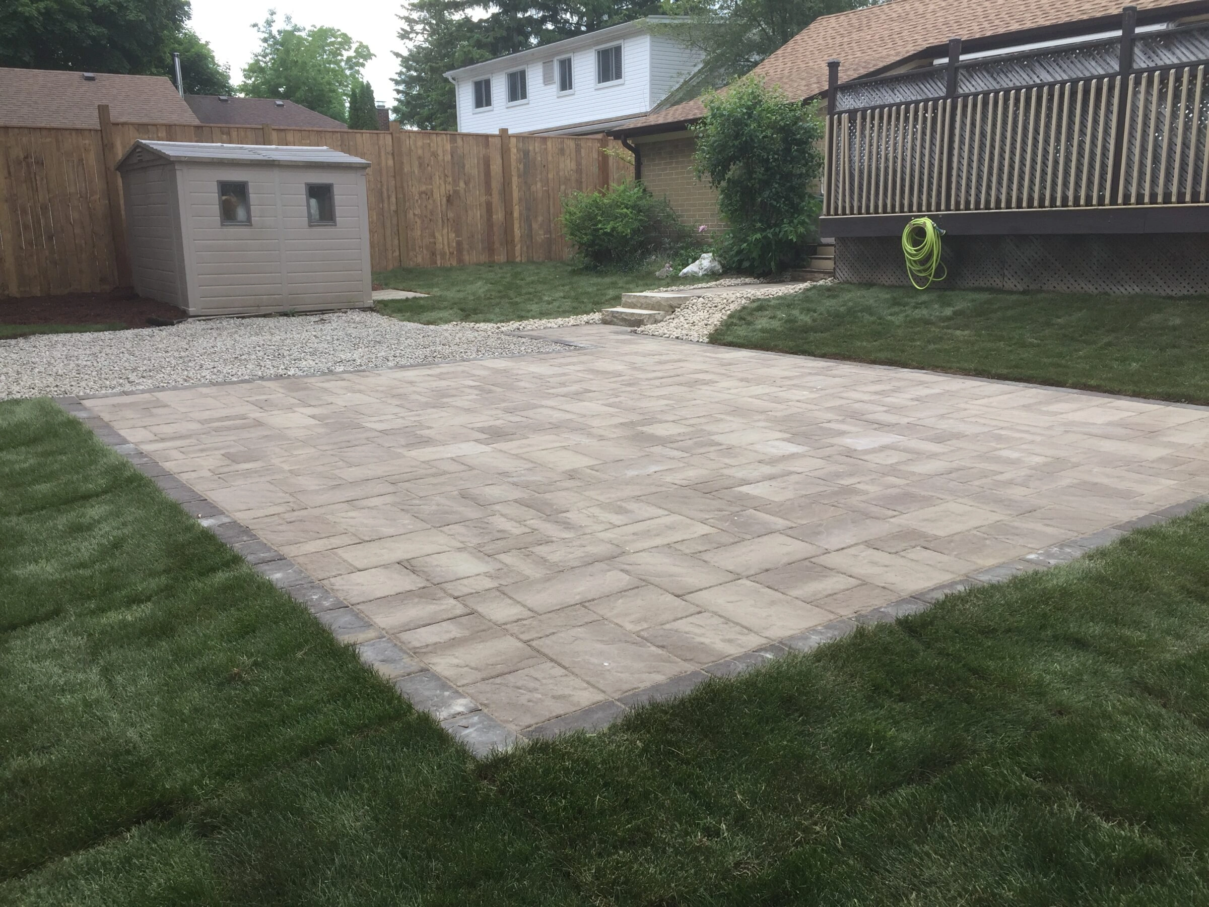 A backyard features a stone patio, grass, wooden fence, small shed, deck with railing, garden hose, and stairs leading up.