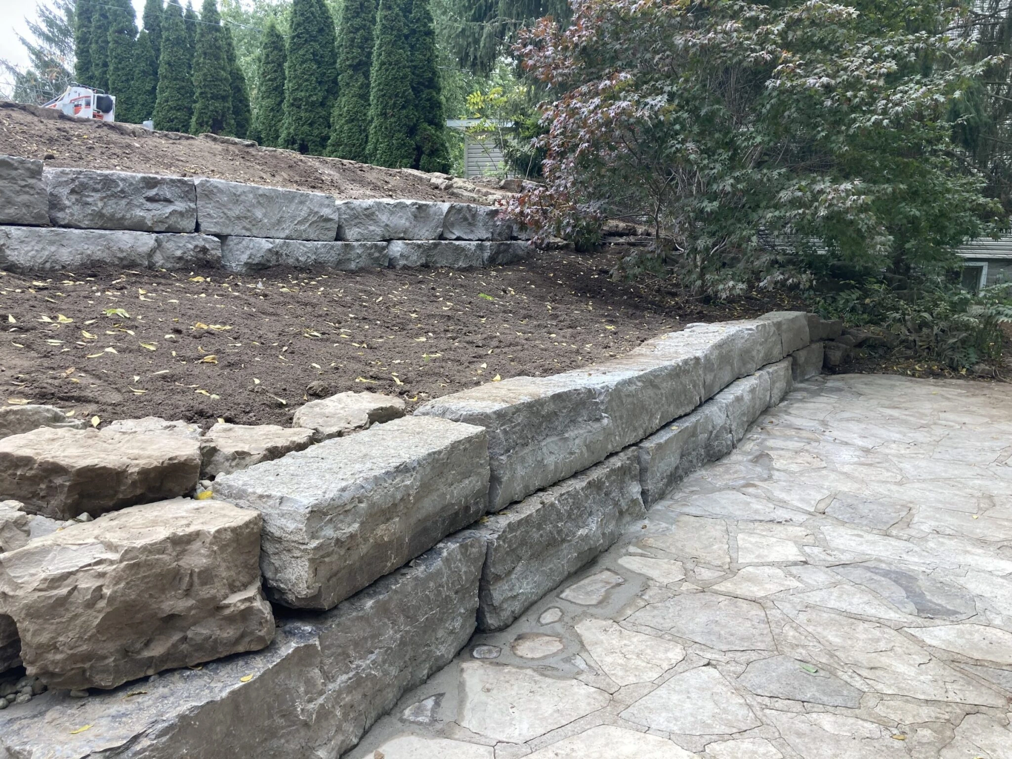Stone retaining wall and patio surrounded by trees, with a freshly dug soil bed and evergreen shrubs in a landscaped outdoor setting.