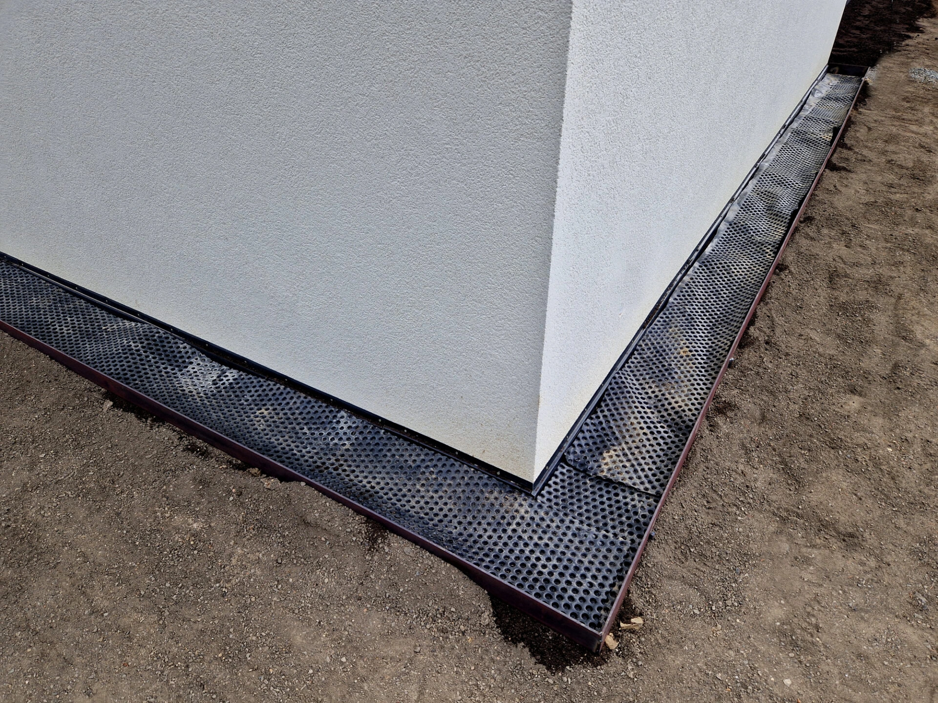 A building's corner with a white wall and perforated black drainage mat surrounded by bare soil, viewed from overhead.