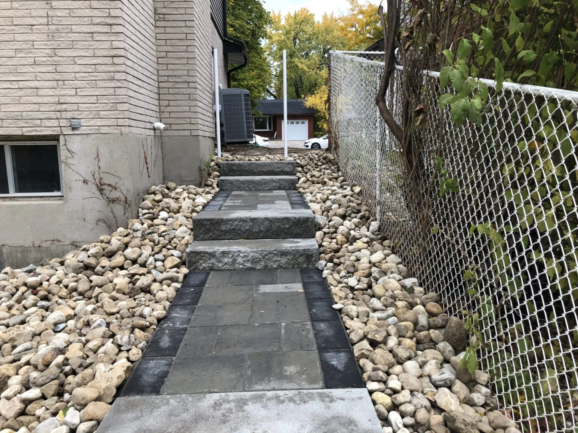 A pathway with stone steps, flanked by rocks, runs alongside a building. A chain-link fence borders greenery. A house is visible ahead.