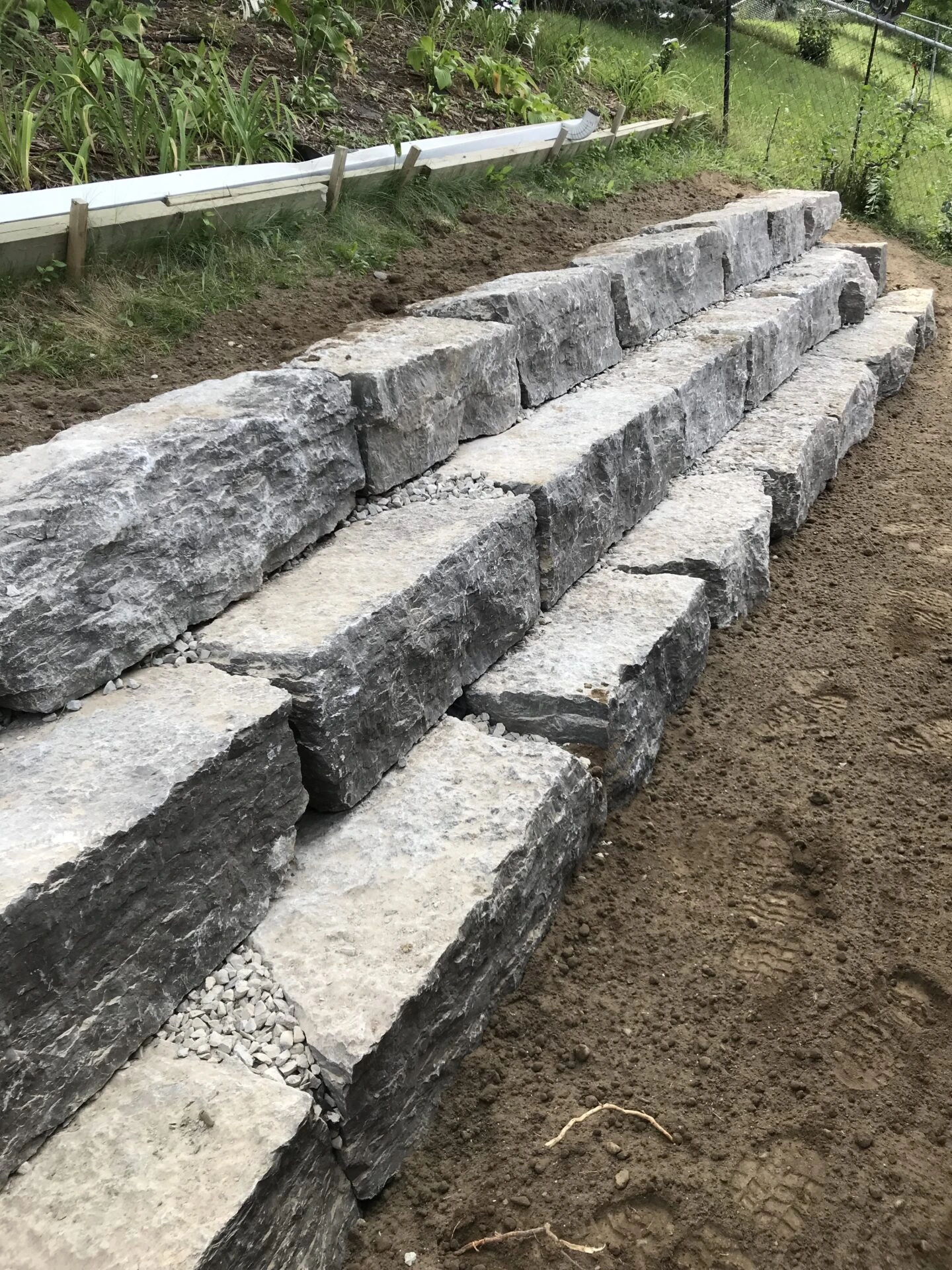 Rough-cut stone blocks form a tiered retaining wall beside a dirt path, bordered by green plants in a garden setting.