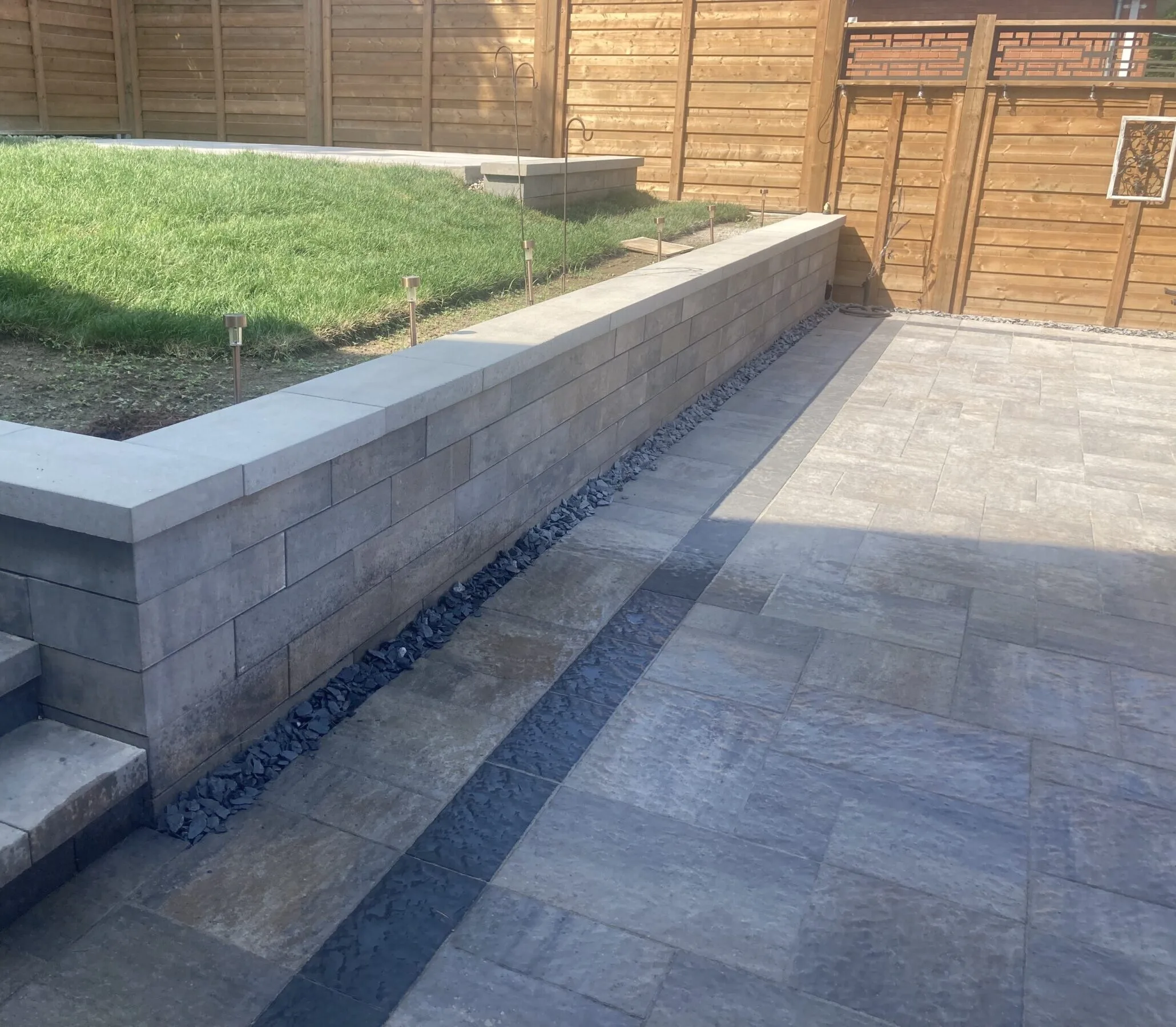 A neatly paved patio with adjacent grassy area, surrounded by a wooden fence, creating a serene outdoor space with modern design elements.