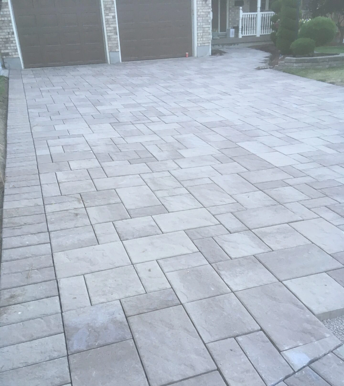 A gray stone driveway with interlocking pavers leads to a garage and porch, featuring tidy, shaped shrubs in front of a brick house.