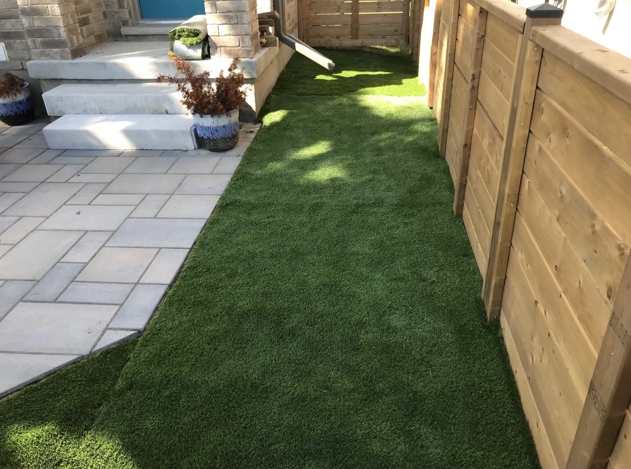 A small backyard with artificial grass, stone steps, and wooden fencing. Two potted plants are placed by the entrance.