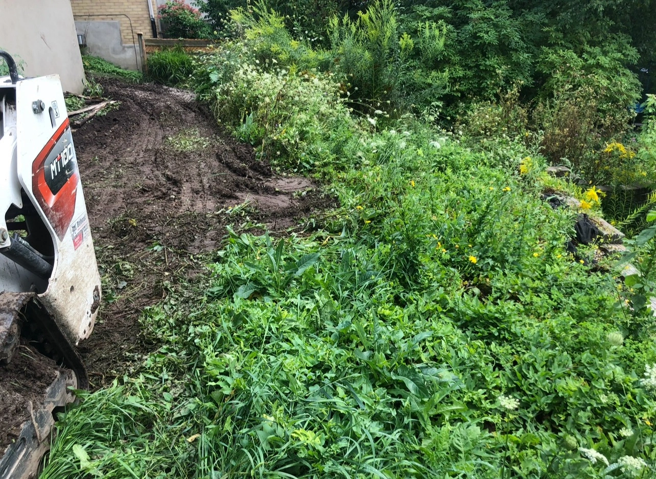 A small construction vehicle is parked on a muddy path next to dense, green vegetation and wildflowers beside a building.