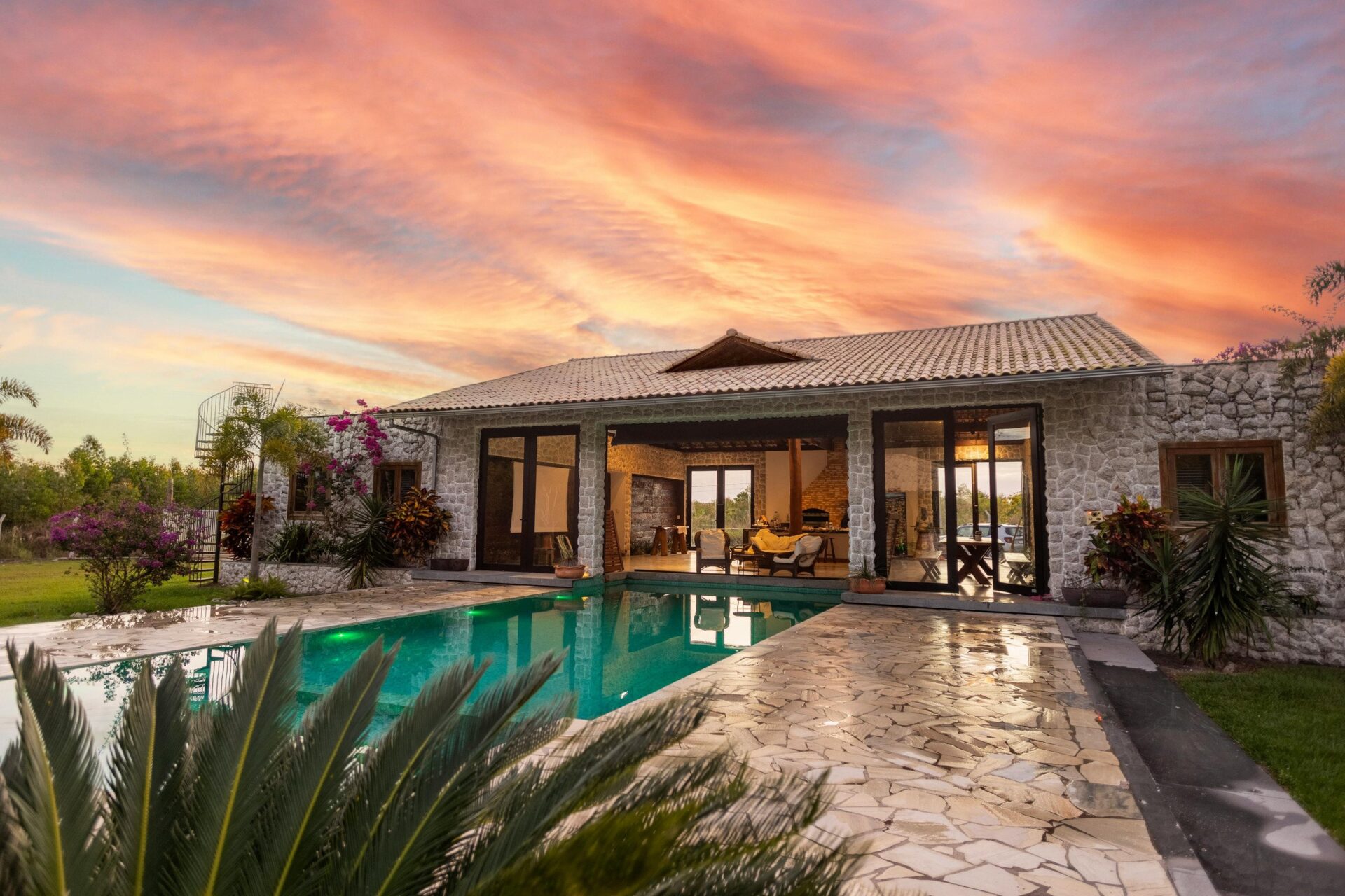 A stone house with large windows, a pool, surrounding plants, and a vibrant sunset sky in a peaceful outdoor setting.