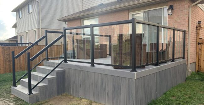 Brick house with modern glass-railed deck, featuring patio furniture and wooden fence. Residential neighborhood setting with grass lawn visible.